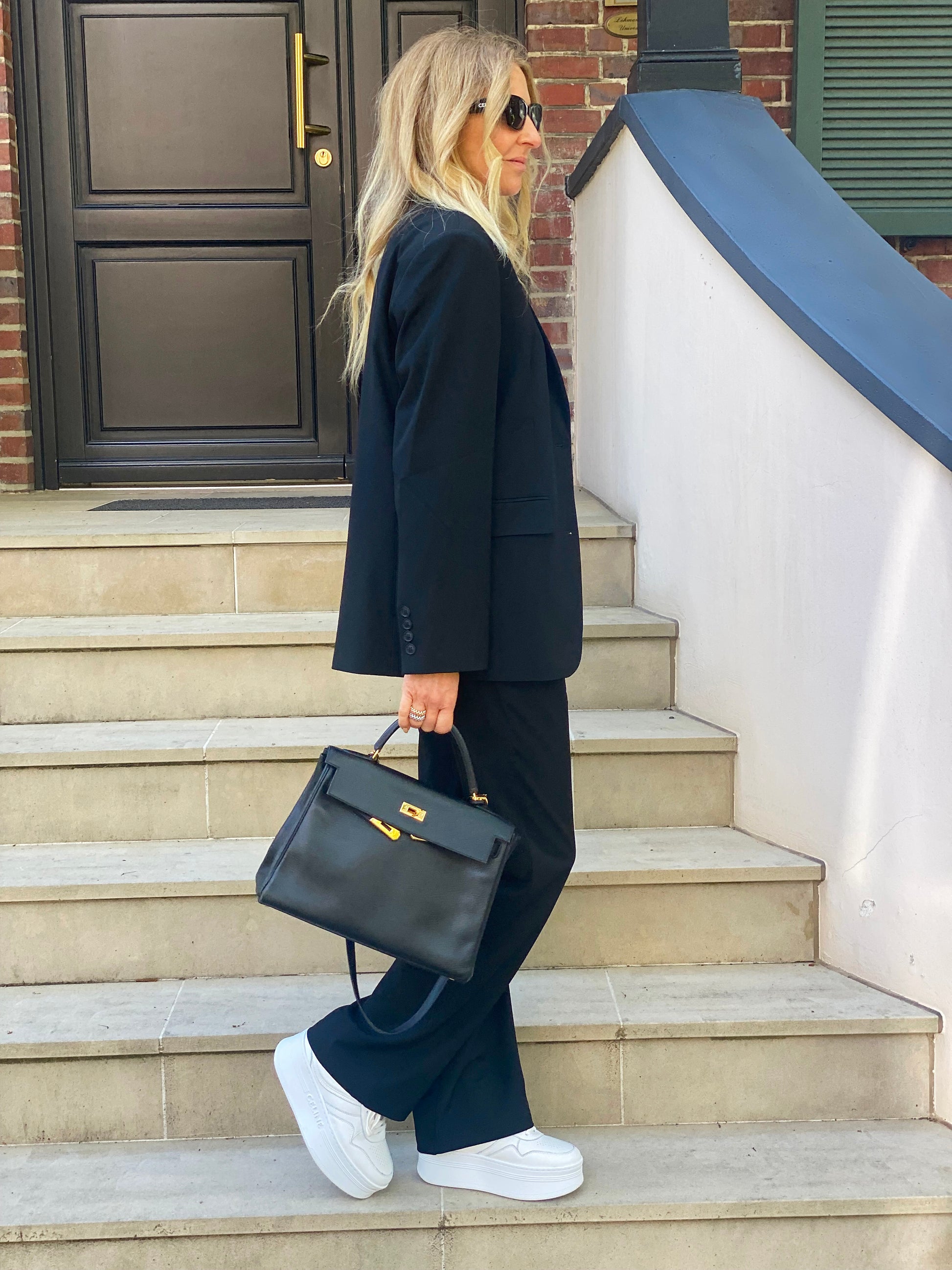 Eine Frau in schicker Geschäftskleidung steht auf einer Freitreppe neben einem Backsteingebäude. Sie trägt eine Hose von Karoline Black zu einem schwarzen Anzug, weiße Turnschuhe, eine Sonnenbrille und eine schwarze Handtasche. Ihr langes blondes Haar fließt, während sie zur Seite blickt.