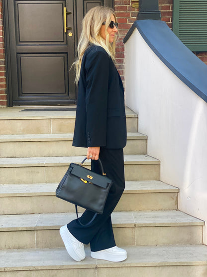 Eine Frau in schicker Geschäftskleidung steht auf einer Freitreppe neben einem Backsteingebäude. Sie trägt eine Hose von Karoline Black zu einem schwarzen Anzug, weiße Turnschuhe, eine Sonnenbrille und eine schwarze Handtasche. Ihr langes blondes Haar fließt, während sie zur Seite blickt.