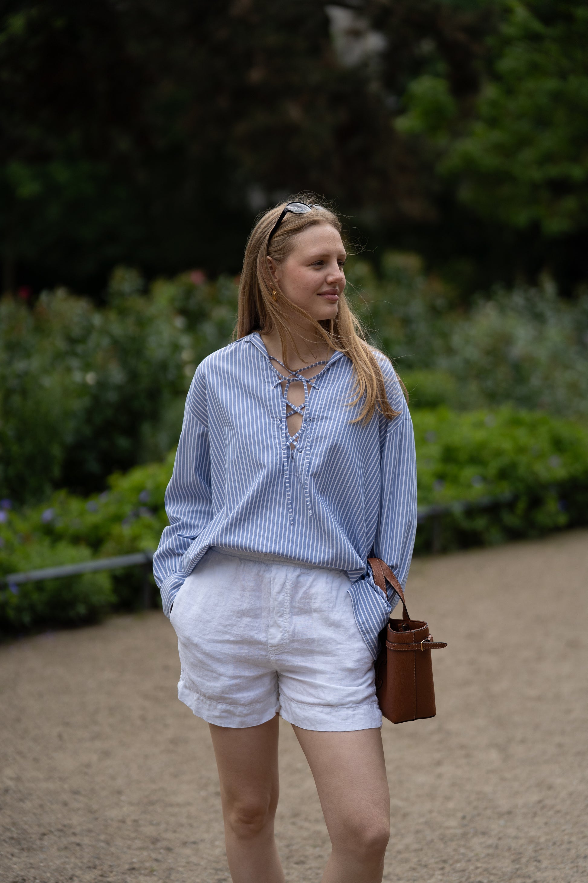 Eine Frau steht im Freien auf einem Kiesweg, trägt die Bluse Fisherman Light Blue Stripes, weiße Shorts und eine Sonnenbrille auf dem Kopf. Sie hält eine braune Tasche in der Hand und ist von Grünzeug umgeben.