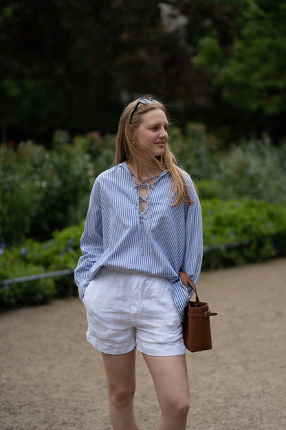 Eine Frau steht im Freien auf einem Kiesweg, trägt die Bluse Fisherman Light Blue Stripes, weiße Shorts und eine Sonnenbrille auf dem Kopf. Sie hält eine braune Tasche in der Hand und ist von Grünzeug umgeben.