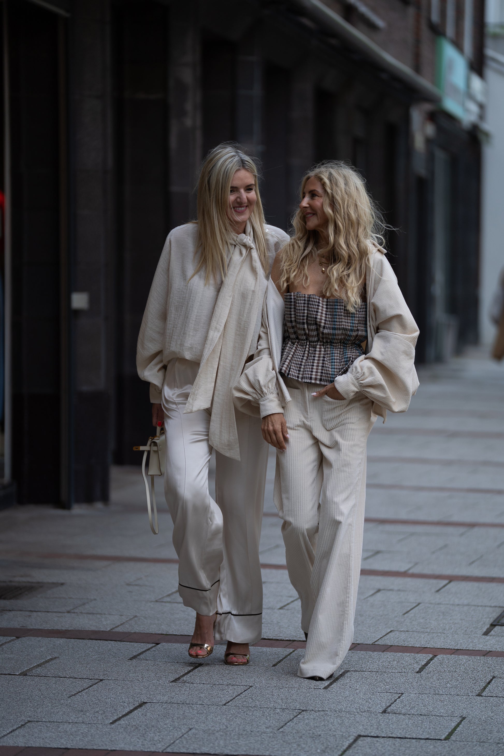Zwei Frauen schlendern Arm in Arm die Stadtstraße entlang, beide lächeln in schicken Cremeweiß-Looks. Ihre Cordhosen 70er Jahre OFW heben sich mit lockeren Oberteilen von den dunklen Gebäuden und dem gepflasterten Bürgersteig ab.