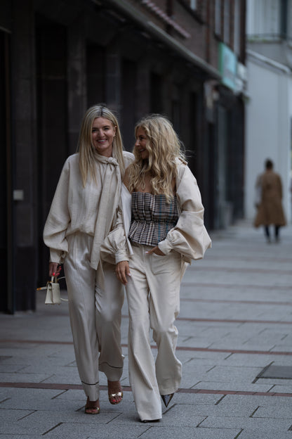 Zwei Frauen gehen zusammen auf einem Bürgersteig in der Stadt spazieren, lächelnd in leichten Outfits. Die eine trägt die Bluse Antonia Corduroy OFW mit einer cremefarbenen Hose, während die andere ein kariertes Oberteil mit einer hellen Cordhose kombiniert.