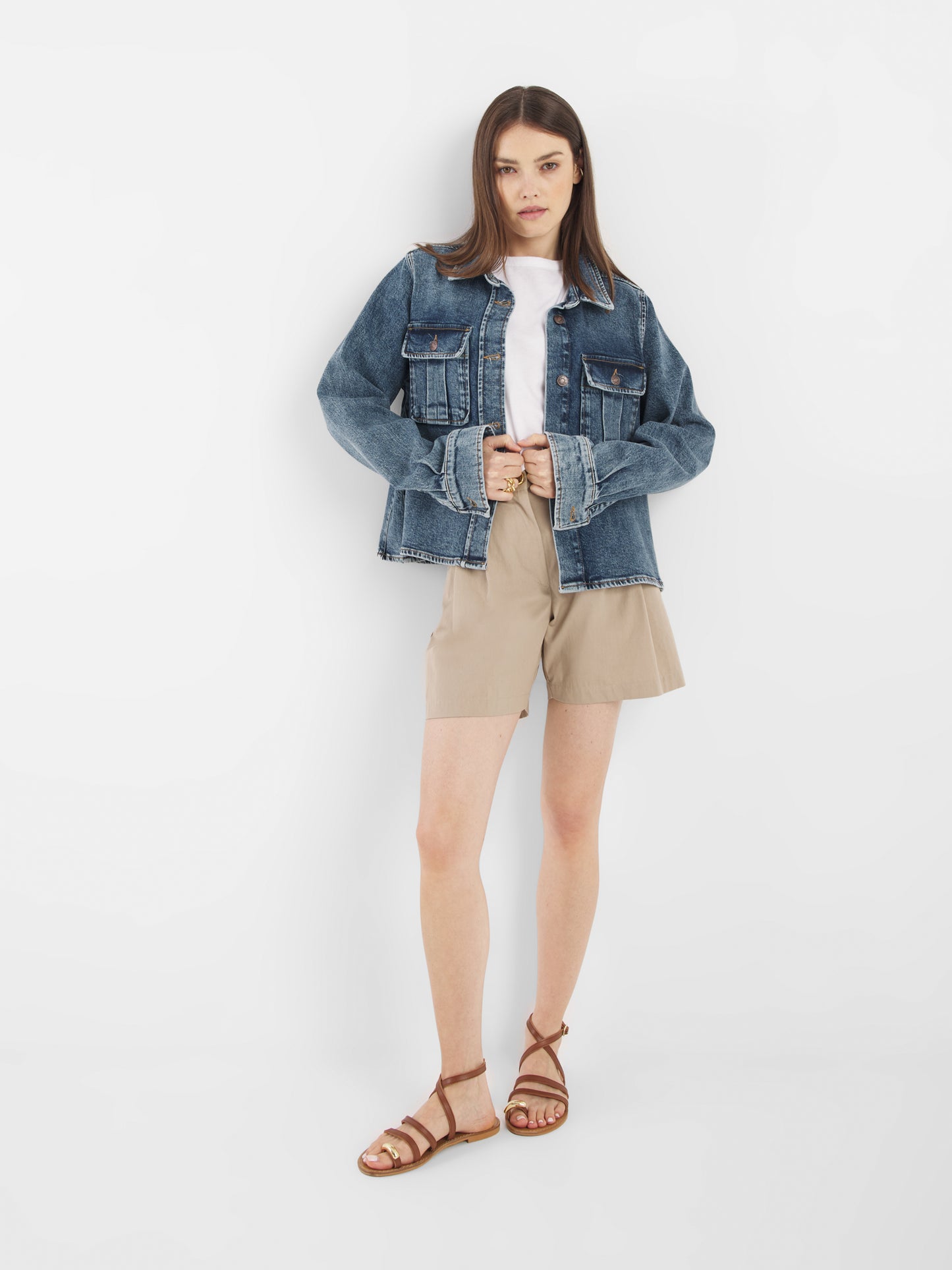 Eine Frau mit glattem Haar und neutralem Gesichtsausdruck steht vor einem weißen Hintergrund und trägt die Jeansjacke Dark Denim über einem weißen Oberteil, beigen Shorts und braunen Riemchensandalen. Sie hält die Vorderseite der Jacke mit beiden Händen fest.