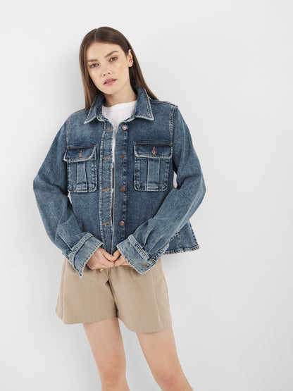 Eine Frau steht vor einer weißen Wand und trägt die Jeansjacke Dark Denim über einem weißen Hemd und beigen Shorts, die Hände in den Jackentaschen und mit neutralem Blick.