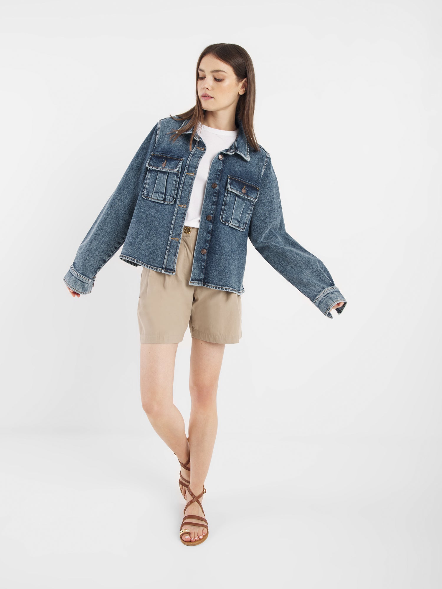 Eine Frau steht vor einem weißen Hintergrund und trägt die Jeansjacke Dark Denim über einem weißen Hemd, beigefarbene Shorts und braune Riemchensandalen. Sie blickt nach unten und hat ein Bein leicht vor dem anderen gekreuzt.