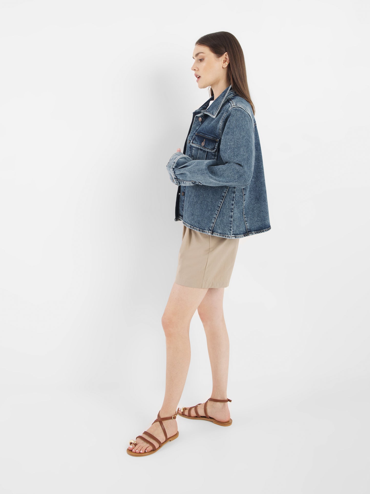 Eine Person steht im Profil vor einem weißen Hintergrund und trägt eine Jeansjacke aus dunklem Denim, beigefarbene Shorts und braune Riemchensandalen.