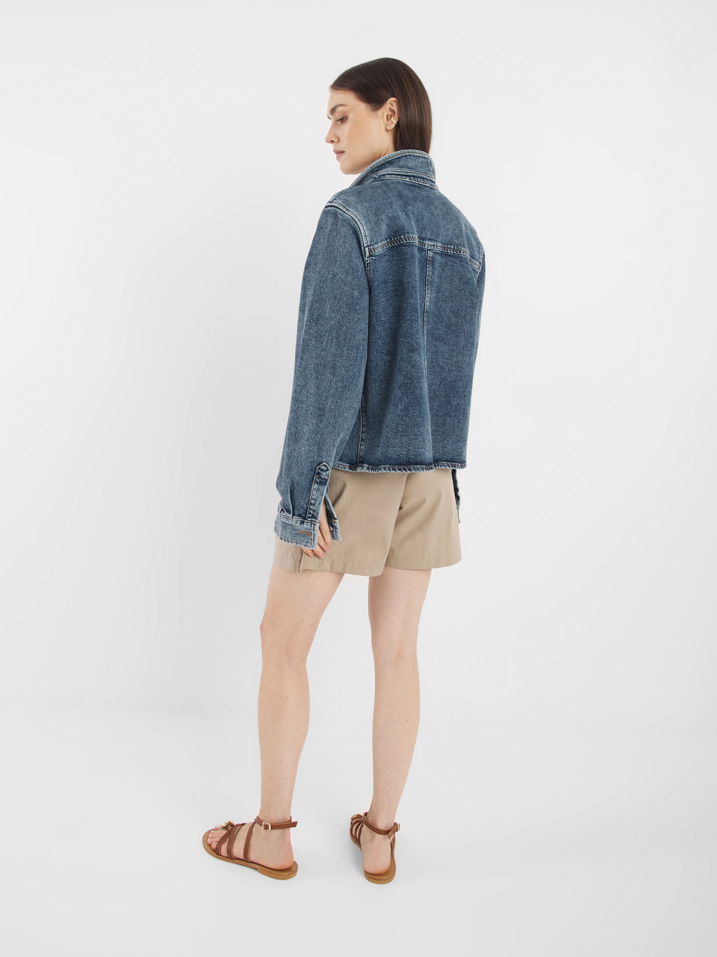 Eine Person steht mit dem Gesicht zur Wand und trägt die Jeansjacke Dark Denim, beige Shorts und braune Sandalen vor einem schlichten weißen Hintergrund.