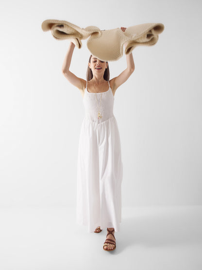 Eine Frau, die das weiße Elisa-Kleid und braune Sandalen trägt, hebt eine gefaltete beigefarbene Decke über ihren Kopf und lächelt vor einem schlichten weißen Hintergrund.