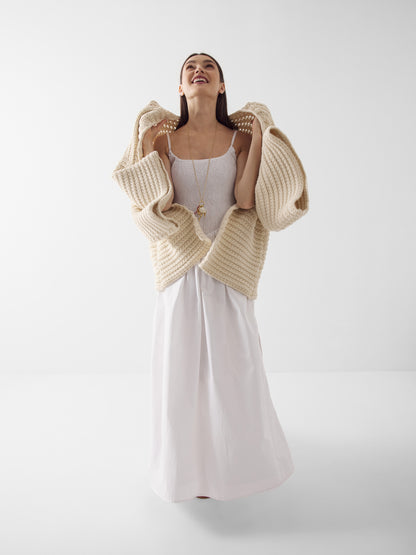 Eine Frau lächelt mit zurückgeneigtem Kopf, sie trägt das Elisa Dress White und eine offene, beigefarbene Strickjacke. Der Hintergrund ist einfarbig weiß.