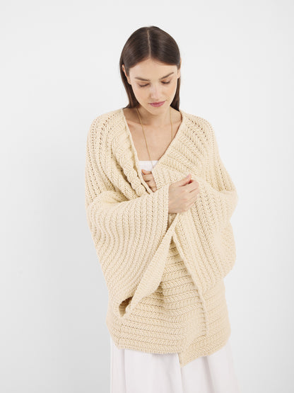 Eine Frau mit glattem braunem Haar trägt die Strickjacke Panna in Einheitsgröße über einem weißen Kleid und hält sie geschlossen, während sie vor einem schlichten weißen Hintergrund nach unten schaut.