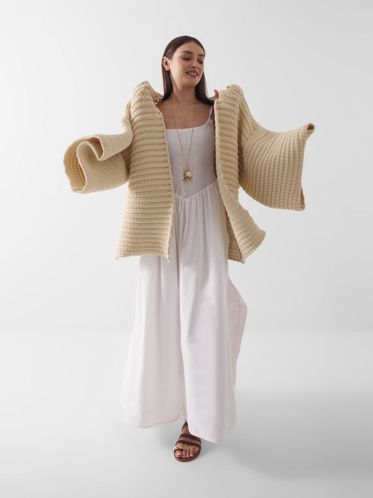 Eine Frau in einem langen weißen Kleid und braunen Sandalen posiert mit dem Cardigan Strick Panna, der über ihre Schultern gelegt ist. Diese dicke beigefarbene Strickjacke sorgt für kuschelige Wärme und Stil, während sie vor einem schlichten weißen Hintergrund lächelt.