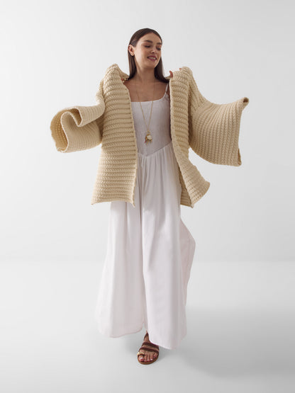 Eine Frau in einem langen weißen Kleid und braunen Sandalen posiert mit dem Cardigan Strick Panna, der über ihre Schultern gelegt ist. Diese dicke beigefarbene Strickjacke sorgt für kuschelige Wärme und Stil, während sie vor einem schlichten weißen Hintergrund lächelt.