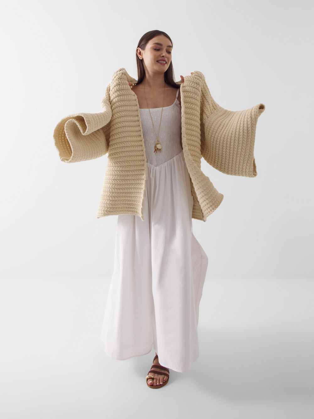 Eine Frau, die das lange weiße Kleid Mirror Mirror On The Wall, hellbraune Sandalen und einen dicken beigen Strickpullover mit weiten Ärmeln trägt, steht vor einem weißen Hintergrund, lächelt mit langen dunklen Haaren und einem leicht erhobenen Arm.