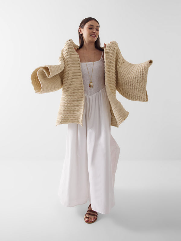 Eine Frau, die das lange weiße Kleid Mirror Mirror On The Wall, hellbraune Sandalen und einen dicken beigen Strickpullover mit weiten Ärmeln trägt, steht vor einem weißen Hintergrund, lächelt mit langen dunklen Haaren und einem leicht erhobenen Arm.