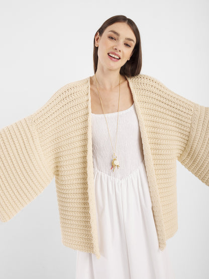 Eine Frau steht mit ausgestreckten Armen und lächelt in dem Cardigan Strick Panna - einer locker sitzenden, cremefarbenen Strickjacke, die über einem weißen Kleid getragen wird. Dieses gemütliche Stück in Einheitsgröße verbindet Komfort mit mühelosem Stil vor einem schlichten weißen Hintergrund.