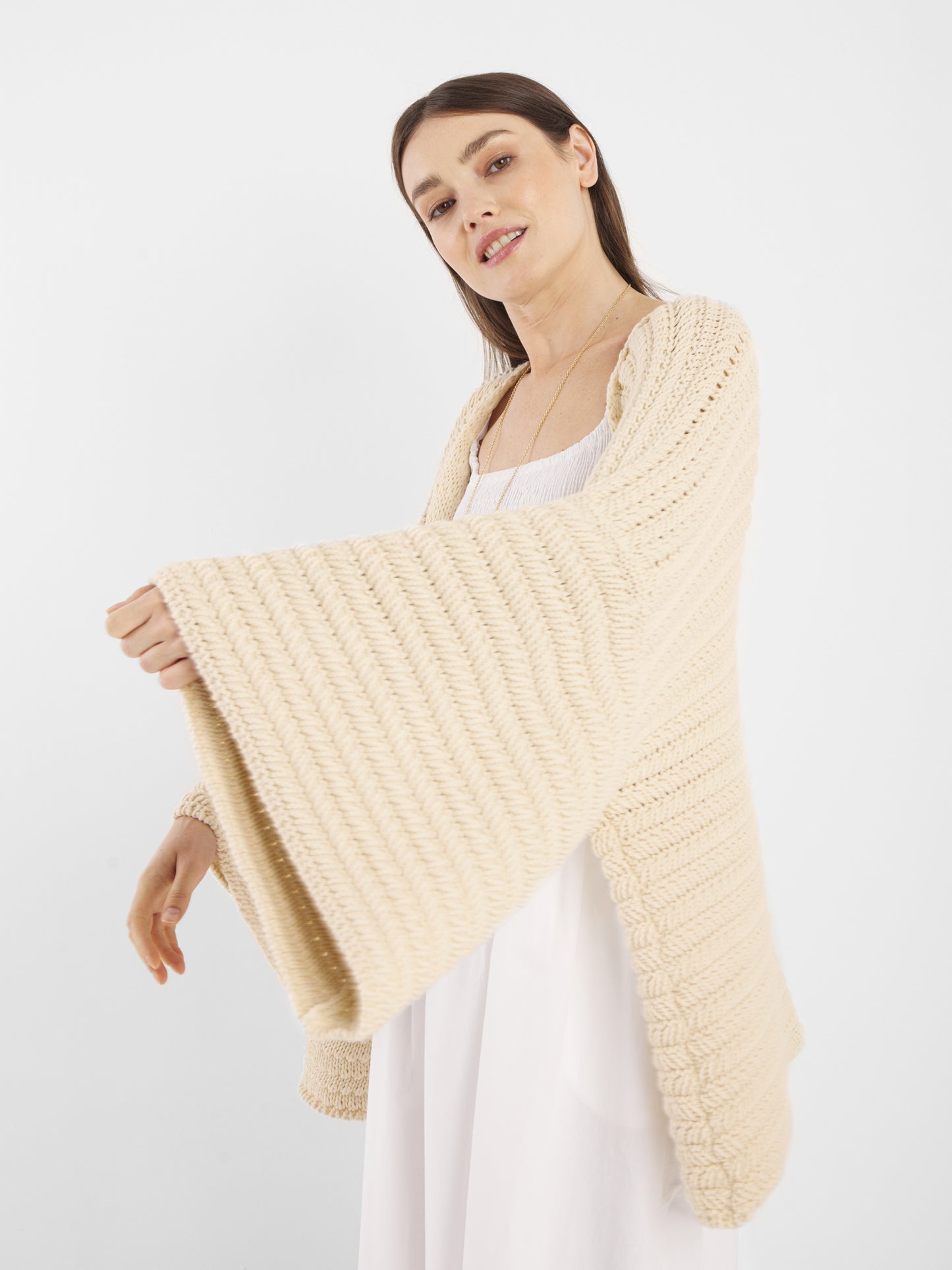 Eine Frau trägt den Cardigan Strick Panna, einen lockeren, cremefarbenen Strick mit weiten Ärmeln, über einem weißen Kleid. Sie steht vor einem schlichten weißen Hintergrund und lächelt leicht. Diese Strickjacke in Einheitsgröße bietet mühelosen Stil und Komfort.