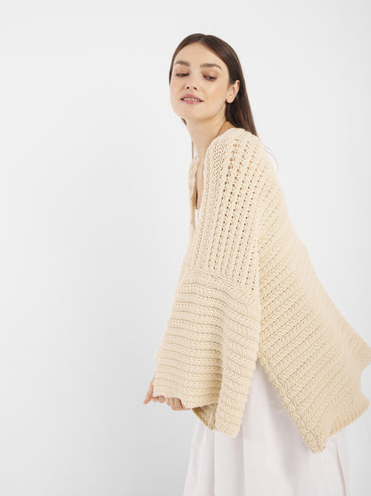 Eine Frau mit langen braunen Haaren lächelt sanft, während sie den Cardigan Strick Panna, eine lockere, cremefarbene Strickjacke in Einheitsgröße über einem weißen Kleid, vor einem schlichten weißen Hintergrund trägt.