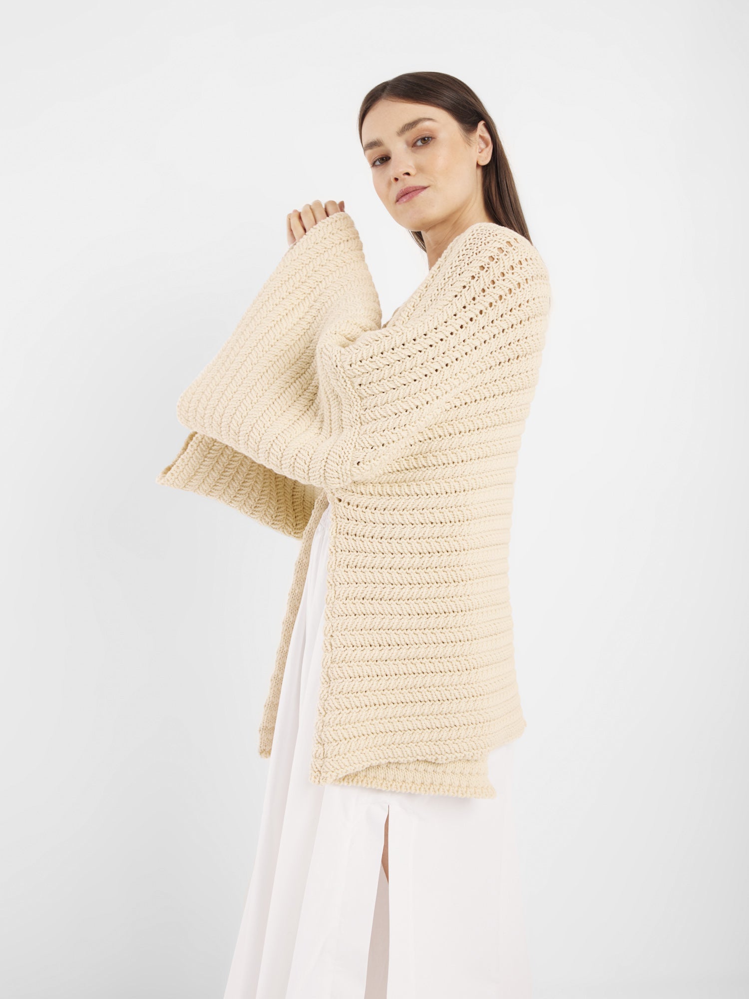 Eine Frau trägt den Cardigan Strick Panna - eine cremefarbene, strukturierte Strickjacke mit weiten Ärmeln über einem weißen Kleid - vor einem schlichten weißen Hintergrund. Dieses modische Kleidungsstück ist in Einheitsgröße erhältlich und bietet vielseitigen Komfort.