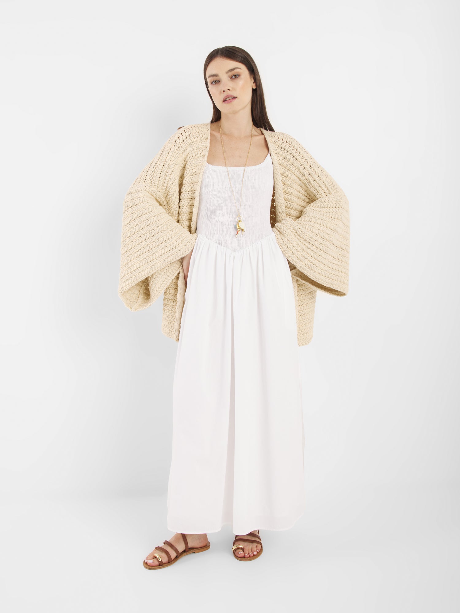 Eine Frau mit glattem braunem Haar und neutralem Gesichtsausdruck steht vor einem weißen Hintergrund und trägt die beigefarbene Strickjacke Strick Panna mit weiten Ärmeln, ein langes weißes Kleid, braune Sandalen und eine lange Halskette.