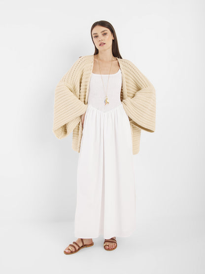 Eine Frau mit glattem braunem Haar und neutralem Gesichtsausdruck steht vor einem weißen Hintergrund und trägt die beigefarbene Strickjacke Strick Panna mit weiten Ärmeln, ein langes weißes Kleid, braune Sandalen und eine lange Halskette.