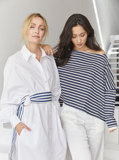 Zwei Frauen stehen in einem Haus. Die eine trägt ein weißes, geknöpftes Kleid mit einem gestreiften Gürtel, die andere das Sweatshirt Sailor Stripe Navy Ecru mit einer weißen Hose. Beide haben einen entspannten Gesichtsausdruck.