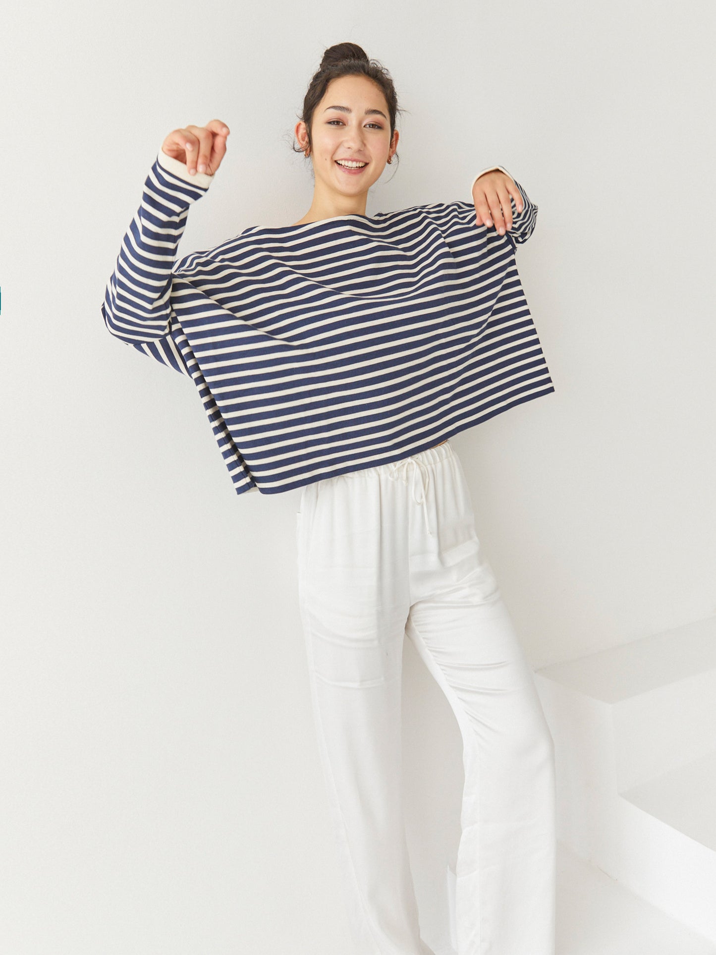 Eine lächelnde Frau steht drinnen vor einem schlichten weißen Hintergrund. Sie trägt das Sweatshirt Sailor Stripe Navy Ecru mit einer lockeren weißen Hose, die Arme angewinkelt und spielerisch erhoben.