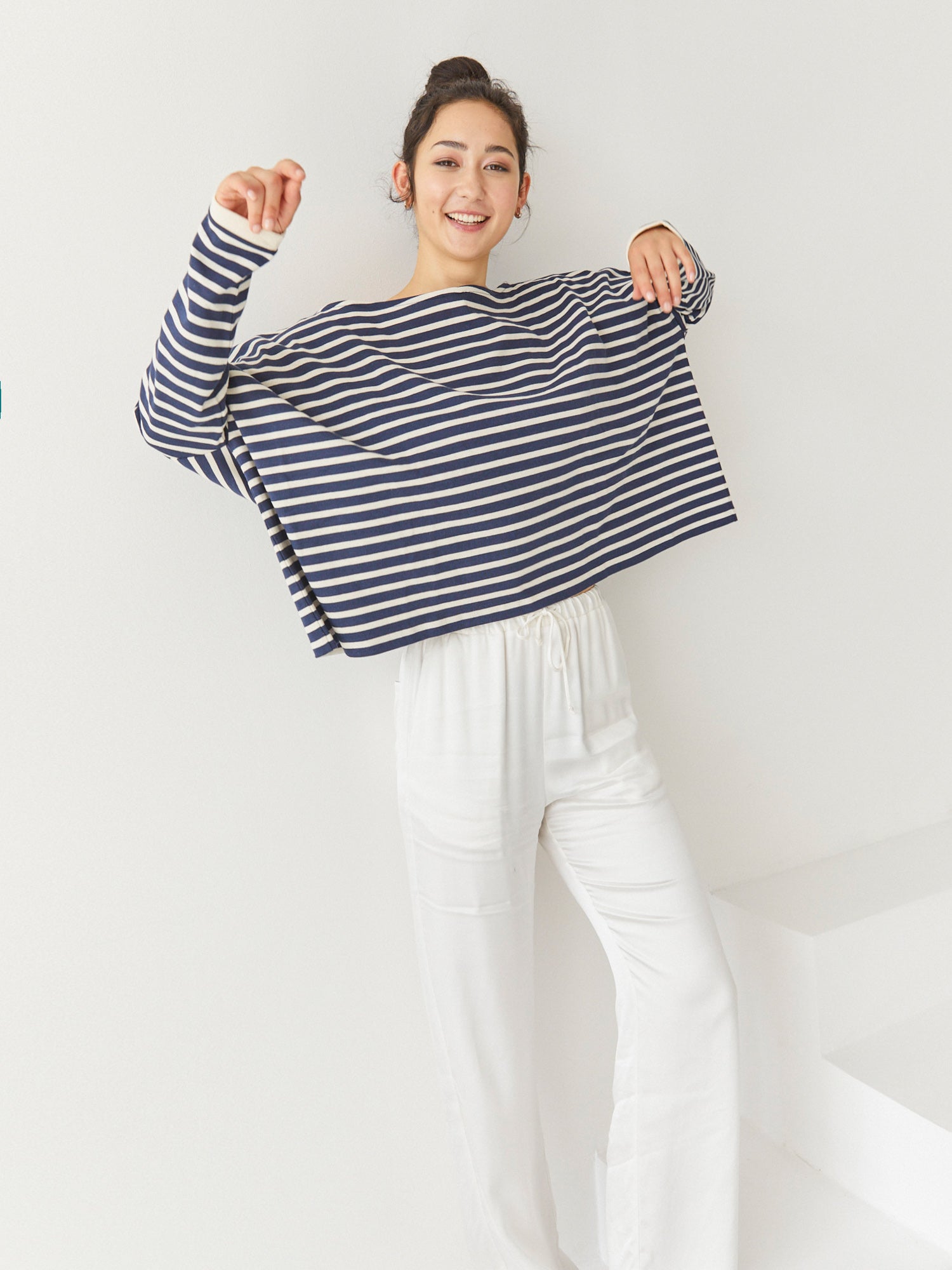 Eine lächelnde Frau steht drinnen vor einem schlichten weißen Hintergrund. Sie trägt das Sweatshirt Sailor Stripe Navy Ecru mit einer lockeren weißen Hose, die Arme angewinkelt und spielerisch erhoben.