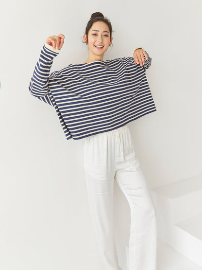 Eine lächelnde Frau steht drinnen vor einem schlichten weißen Hintergrund. Sie trägt das Sweatshirt Sailor Stripe Navy Ecru mit einer lockeren weißen Hose, die Arme angewinkelt und spielerisch erhoben.