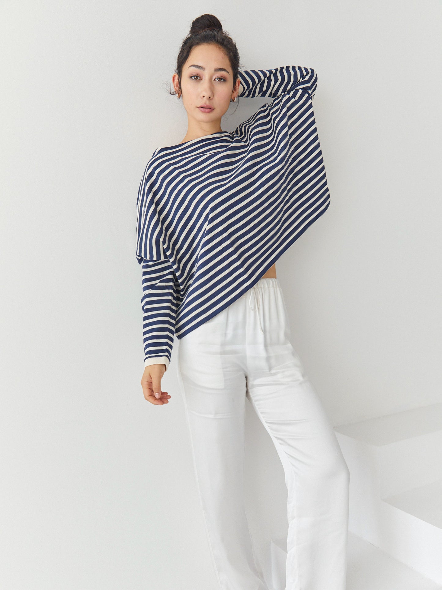 Eine Frau steht in einem Innenraum vor einer weißen Wand. Sie trägt das Sweatshirt Sailor Stripe Navy Ecru mit klassischen bretonischen Streifen und eine weiße Hose. Ihr Haar ist hochgesteckt, und sie hat einen Arm erhoben, der auf ihrem Kopf ruht.