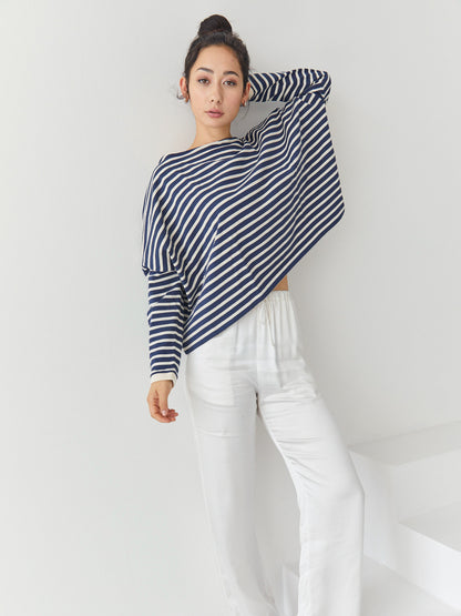 Eine Frau steht in einem Innenraum vor einer weißen Wand. Sie trägt das Sweatshirt Sailor Stripe Navy Ecru mit klassischen bretonischen Streifen und eine weiße Hose. Ihr Haar ist hochgesteckt, und sie hat einen Arm erhoben, der auf ihrem Kopf ruht.
