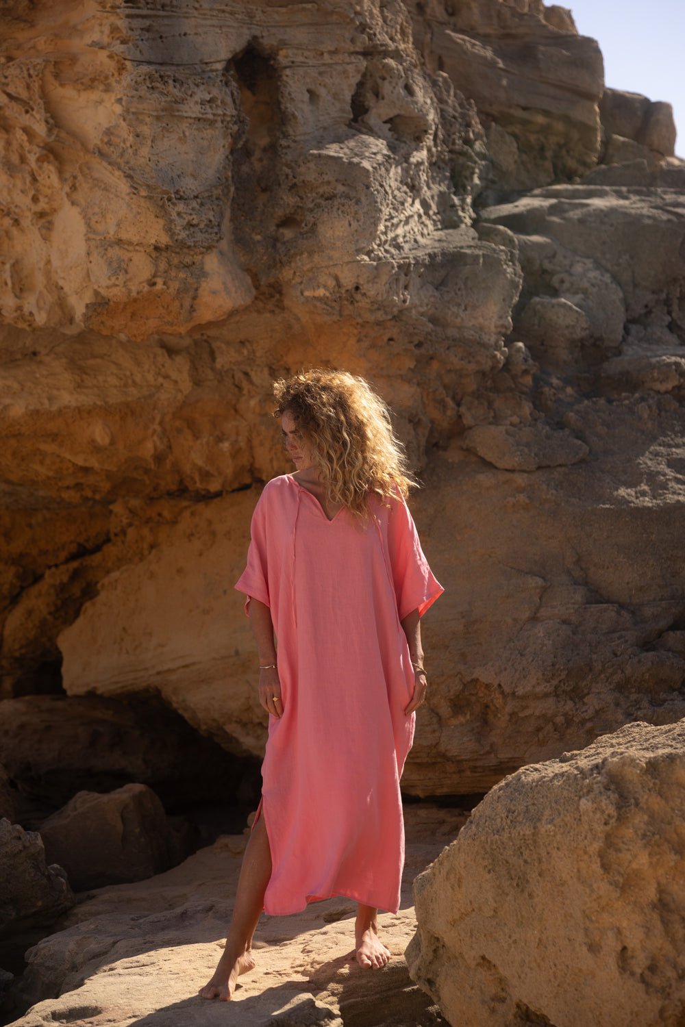 Eine Person mit lockigem Haar trägt das lockere rosa Let's Go Marrakesch Kaftan Kleid und steht barfuß auf einem felsigen Gelände mit großen Steinformationen und Sonnenlicht, das im Hintergrund warme Schatten wirft.