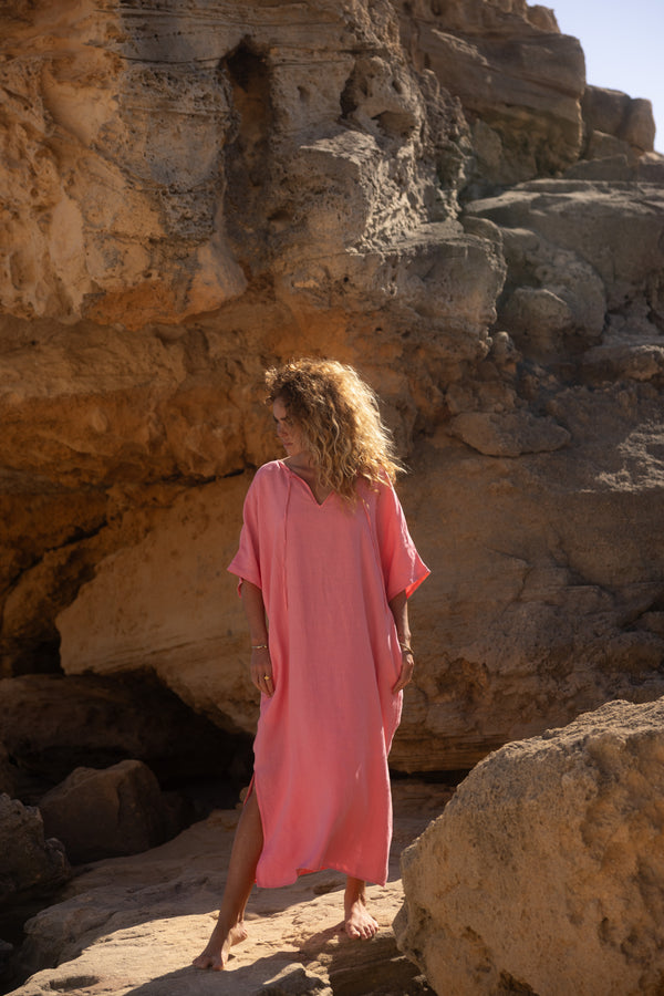 Eine Person mit lockigem Haar trägt das lockere rosa Let's Go Marrakesch Kaftan Kleid und steht barfuß auf einem felsigen Gelände mit großen Steinformationen und Sonnenlicht, das im Hintergrund warme Schatten wirft.