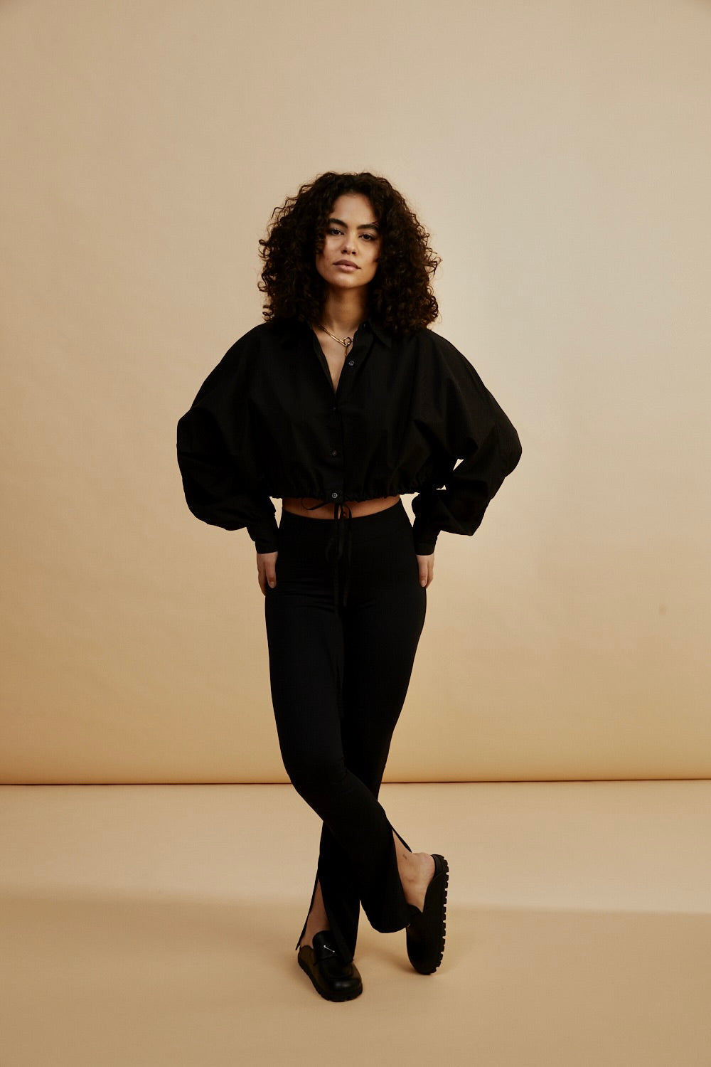 Eine Frau mit lockigem Haar posiert vor einem beigen Hintergrund und trägt eine schwarze Kurzjacke und geschlitzte Leggings - 80% Recycled. Sie steht mit den Händen hinter dem Rücken und gekreuzten Knöcheln.