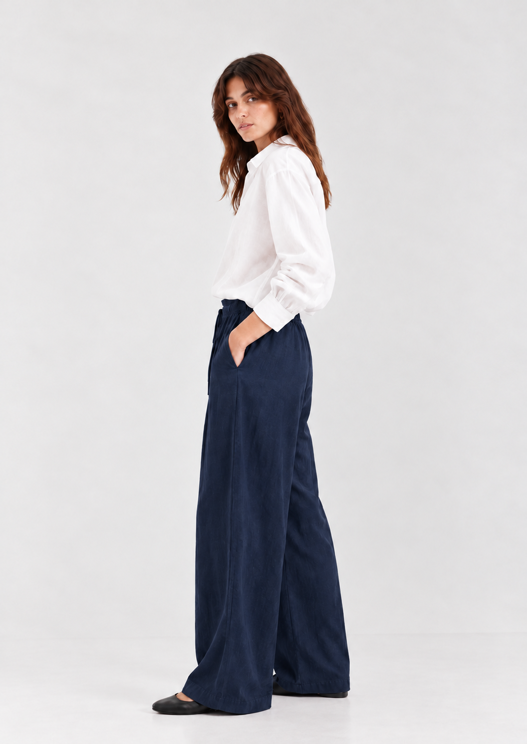 Eine Frau in einem weißen, langärmeligen Hemd mit Knöpfen und einer locker sitzenden Hose Josy Linen Navy, die die Hände in den Taschen hat.