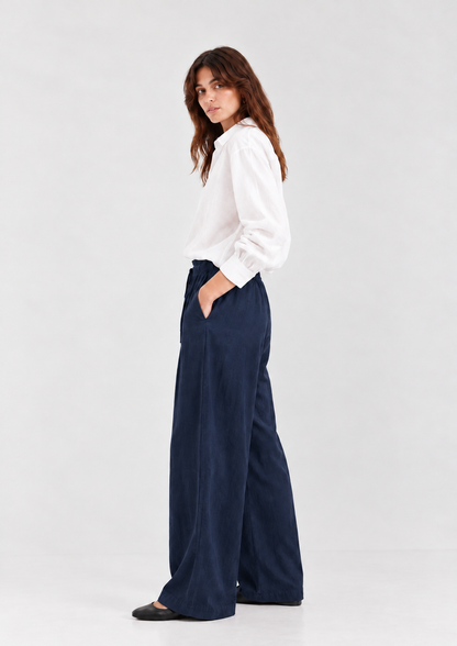 Eine Frau in einem weißen, langärmeligen Hemd mit Knöpfen und einer locker sitzenden Hose Josy Linen Navy, die die Hände in den Taschen hat.