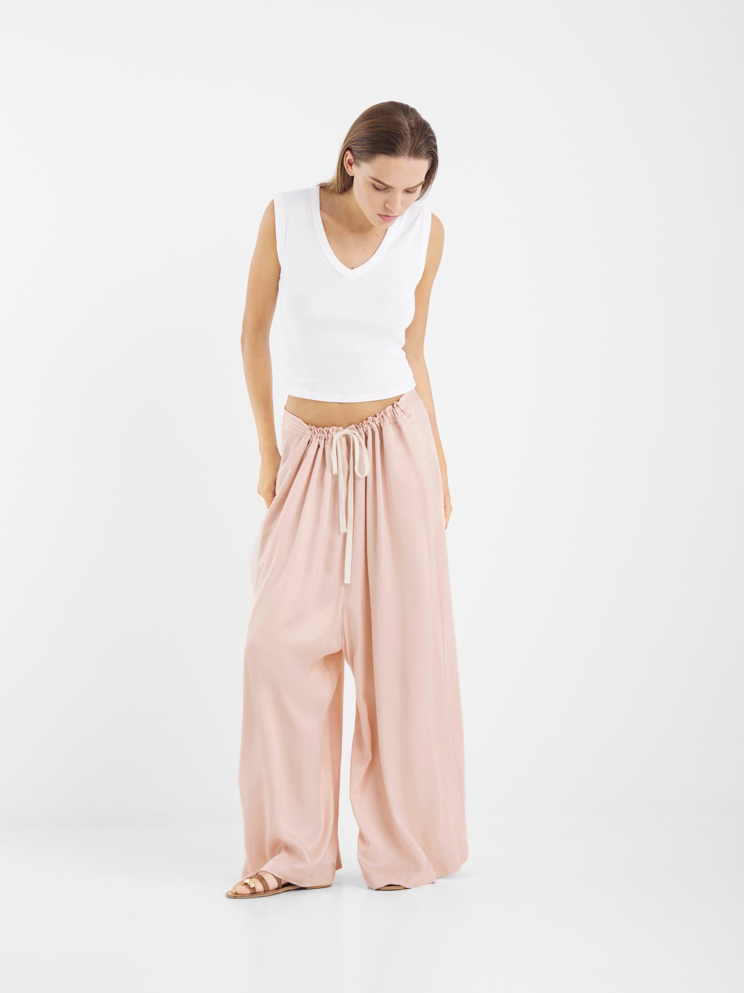 Eine Frau steht vor einem einfarbig weißen Hintergrund und trägt ein ärmelloses weißes Oberteil mit V-Ausschnitt, eine Hose in Übergröße in Rot mit Kordelzug und beigefarbene Sandalen. Sie blickt nach unten und hat die Hände in den Taschen.