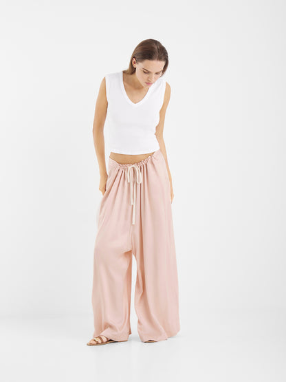 Eine Frau steht vor einem einfarbig weißen Hintergrund und trägt ein ärmelloses weißes Oberteil mit V-Ausschnitt, eine Hose in Übergröße in Rot mit Kordelzug und beigefarbene Sandalen. Sie blickt nach unten und hat die Hände in den Taschen.