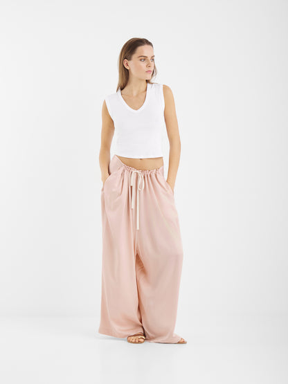 Eine Frau posiert vor einem weißen Hintergrund und trägt ein ärmelloses weißes Crop-Top und Pants Oversize - eine weite, hochtaillierte Hose in Rot mit weitem Bein und Kordelzug in der Taille. Sie hat die Hände in den Taschen und blickt zur Seite.