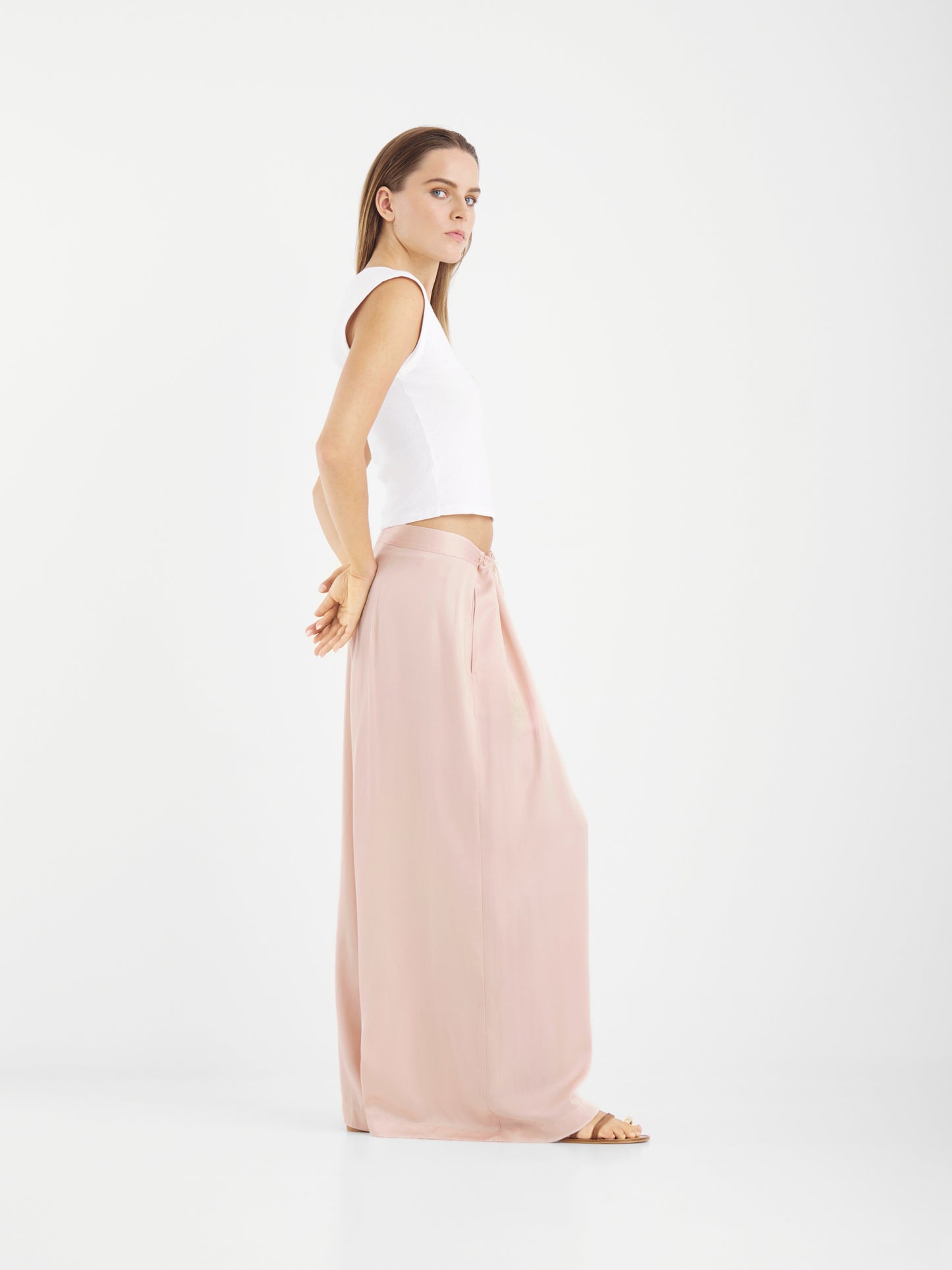 Eine Frau steht im Profil vor einem weißen Hintergrund. Sie trägt ein weißes ärmelloses Oberteil und eine lockere, errötete Hose in Übergröße. Ihre Arme sind entspannt hinter ihrem Rücken verschränkt, während sie mit neutralem Blick nach vorne blickt.