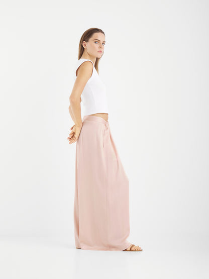 Eine Frau steht im Profil vor einem weißen Hintergrund. Sie trägt ein weißes ärmelloses Oberteil und eine lockere, errötete Hose in Übergröße. Ihre Arme sind entspannt hinter ihrem Rücken verschränkt, während sie mit neutralem Blick nach vorne blickt.