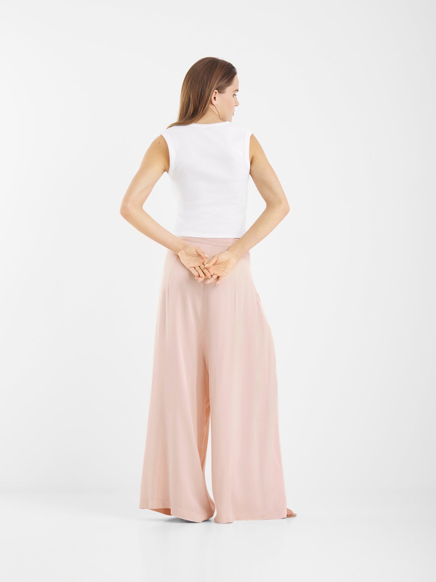 Eine Frau steht mit dem Gesicht nach hinten, die Hände hinter dem Rücken verschränkt, und trägt ein ärmelloses weißes Oberteil und eine Hose in Übergröße mit weitem Bein und Satinoptik. Der Hintergrund ist einfarbig weiß.