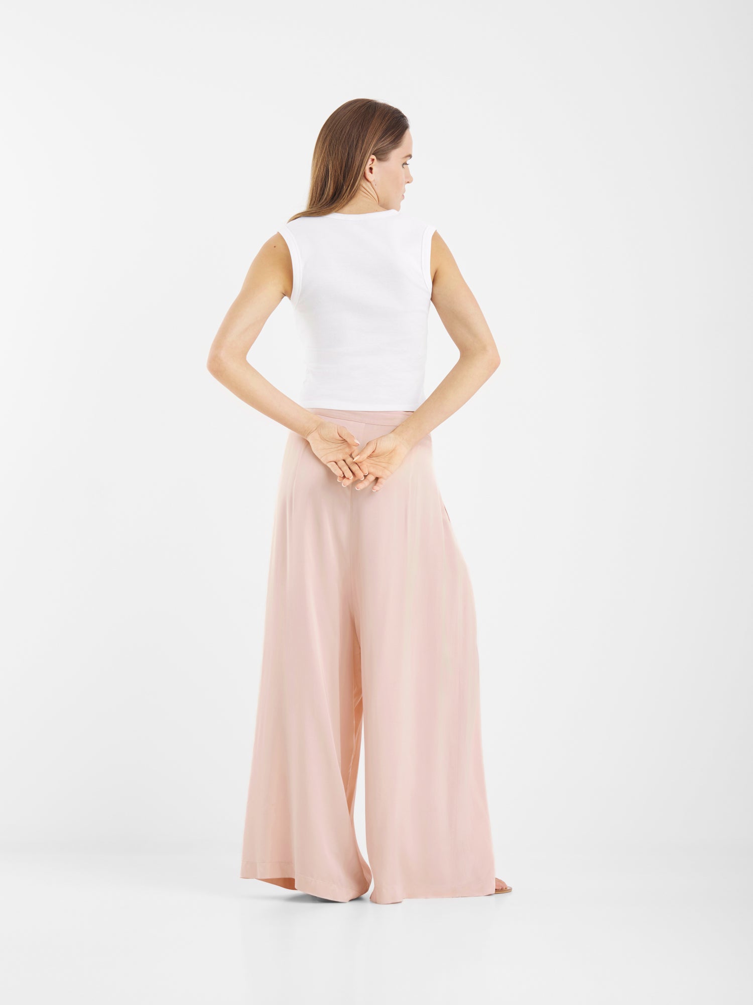 Eine Frau steht mit dem Gesicht nach hinten, die Hände hinter dem Rücken verschränkt, und trägt ein ärmelloses weißes Oberteil und eine Hose in Übergröße mit weitem Bein und Satinoptik. Der Hintergrund ist einfarbig weiß.