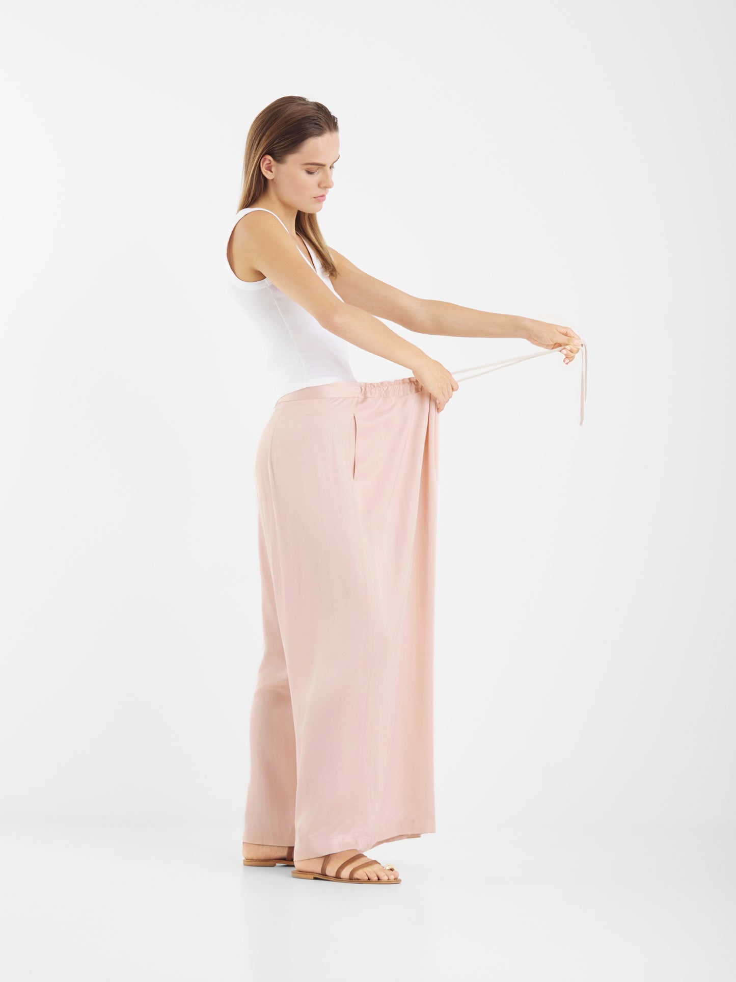 Eine Frau in einem weißen Tank-Top und einer lockeren, erröteten Oversize-Hose steht seitlich und hält den Bund heraus, um die geräumige Passform zu zeigen. Sie trägt flache Sandalen und posiert entspannt vor einem schlichten weißen Hintergrund.