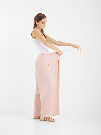 Eine Frau in einem weißen Tank-Top und einer lockeren, erröteten Oversize-Hose steht seitlich und hält den Bund heraus, um die geräumige Passform zu zeigen. Sie trägt flache Sandalen und posiert entspannt vor einem schlichten weißen Hintergrund.