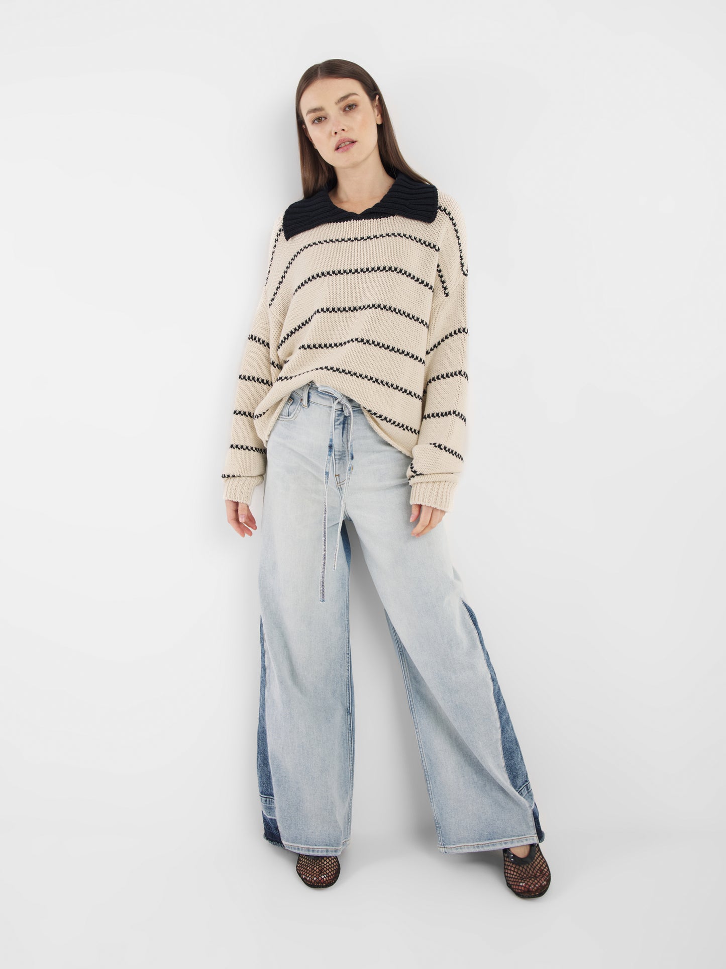 Eine Frau mit glattem braunem Haar steht vor einem weißen Hintergrund. Sie trägt den Pullover Polo Ecru Dark Blue und hellblaue Jeans mit weitem Bein und dunkelblauen Einsätzen, dazu schwarze Netzschuhe.