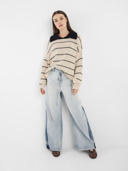 Eine Frau mit glattem braunem Haar steht vor einem weißen Hintergrund. Sie trägt den Pullover Polo Ecru Dark Blue und hellblaue Jeans mit weitem Bein und dunkelblauen Einsätzen, dazu schwarze Netzschuhe.