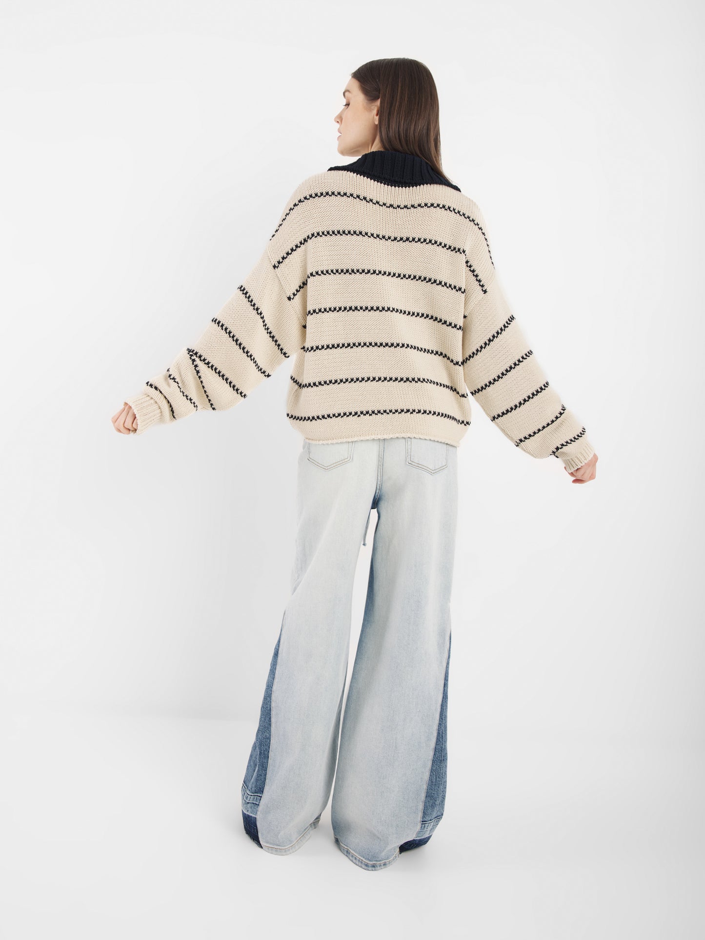 Eine Person steht vor einem weißen Hintergrund und trägt den Pullover Polo Ecru Dark Blue - einen cremefarbenen und schwarz gestreiften Pullover - und hellblaue Jeans mit weitem Bein und dunklen Jeanseinsätzen.