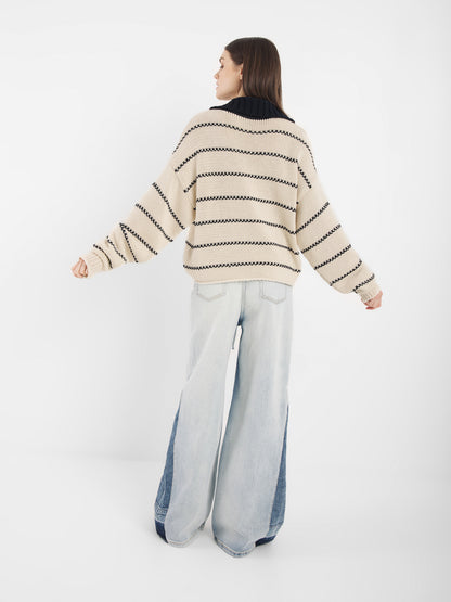 Eine Person steht vor einem weißen Hintergrund und trägt den Pullover Polo Ecru Dark Blue - einen cremefarbenen und schwarz gestreiften Pullover - und hellblaue Jeans mit weitem Bein und dunklen Jeanseinsätzen.