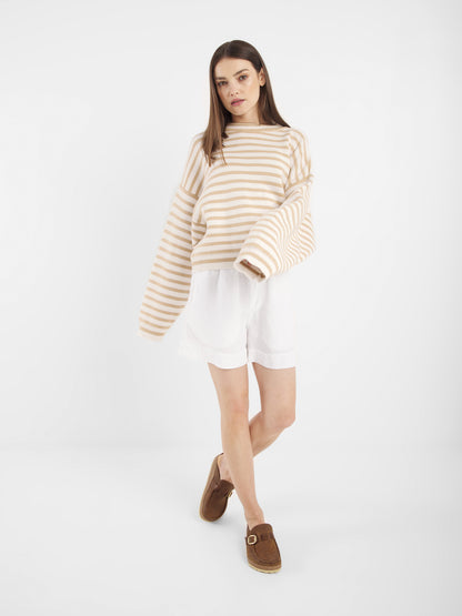 Eine Frau mit glattem braunem Haar und neutralem Gesichtsausdruck steht vor einem weißen Hintergrund und trägt den Pullover Sailor Beige Offwhite, gepaart mit weißen Shorts und braunen Slippern.