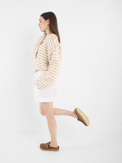 Eine Frau modelliert den Pullover Sailor Beige Offwhite, gepaart mit weißen Shorts und braunen Slippern, seitlich stehend mit einem angewinkelten Bein vor einem schlichten weißen Hintergrund.